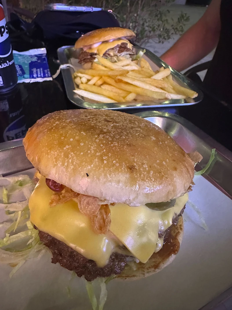 A close-up of a cheeseburger on a metal tray at a restaurant in Krabi. Another plate with a burger and fries is visible in the background.