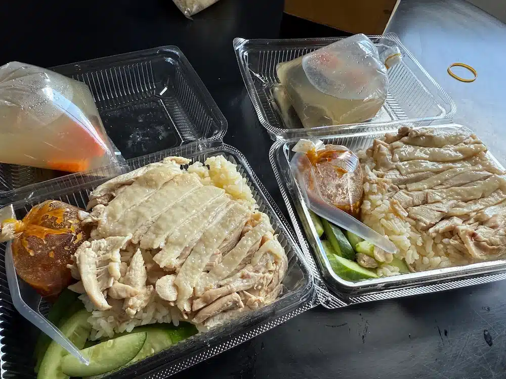 A takeaway foam box filled with tender steamed chicken over oily rice, accompanied by a bag of hot soup and spicy dipping sauce.
