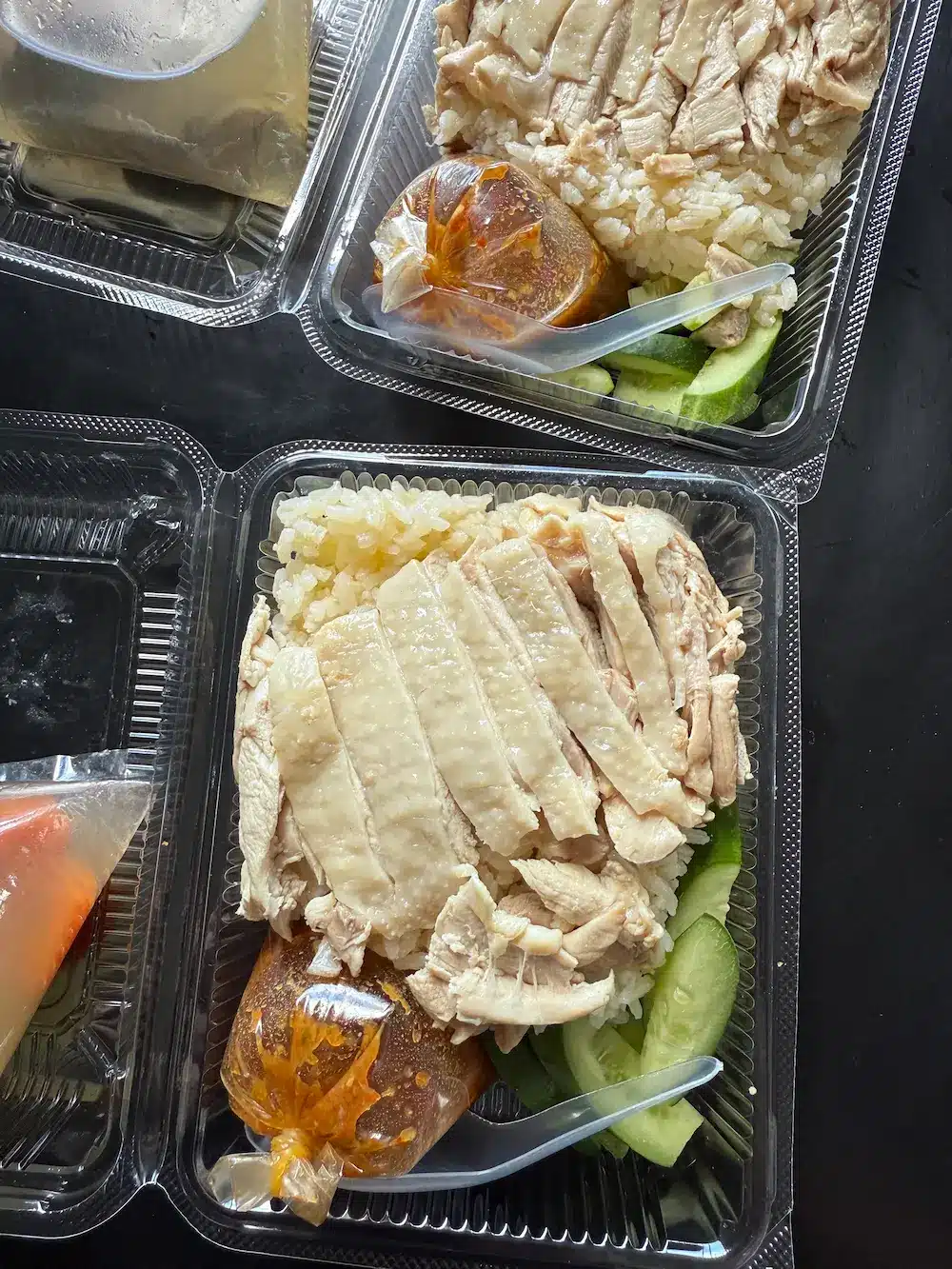 A takeaway foam box filled with tender steamed chicken over oily rice, accompanied by a bag of hot soup and spicy dipping sauce.