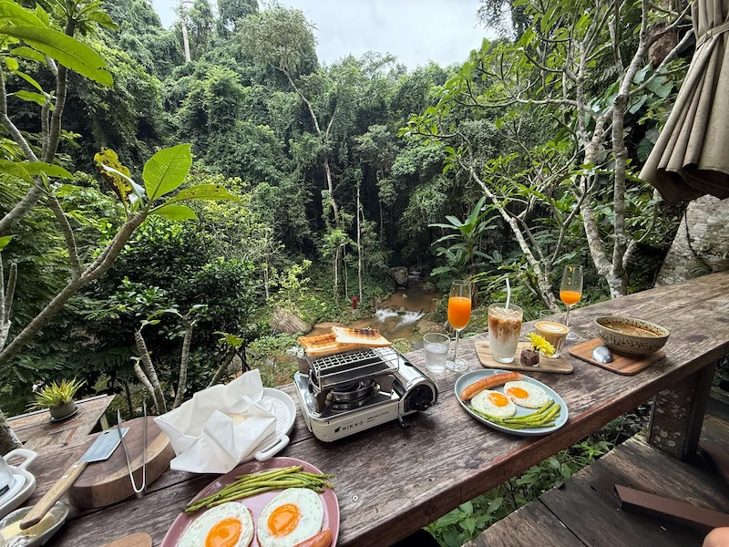 A delicious breakfast spread at Woo Ma Ca Moo featuring eggs, toast, and coffee, set on a wooden balcony overlooking a lush green jungle and stream.