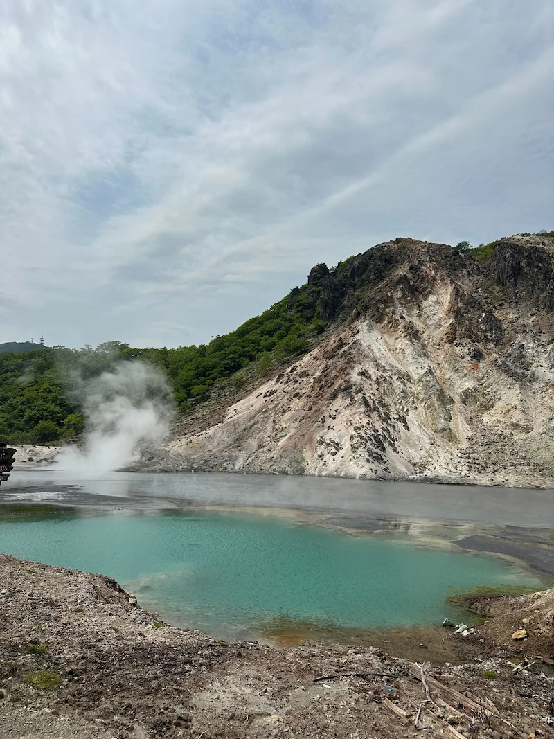 The turquoise waters of a volcanic pond at noboribetsu jigokudani valley with active steam vents rising from the rocky mountain slope.