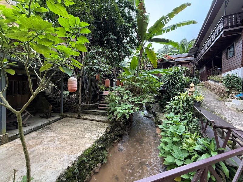 A wooden walkway and small bridge crossing over a natural stream, surrounded by traditional wooden buildings.