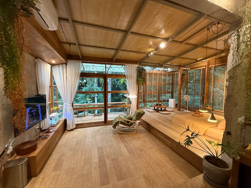 The spacious interior of the Woo Ma Ca Moo villa featuring a large bed on a raised platform, floor-to-ceiling windows, and a relaxing rocking chair.