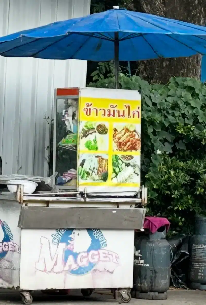 A vertical close-up of the street vendor's cart featuring a bright yellow sign with red Thai script reading "Khao Man Gai" (Chicken Rice).