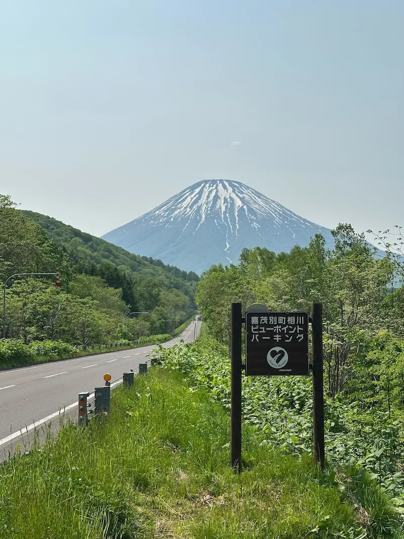A picturesque road stretching towards the majestic Mt. Yotei, marked by a "Scenic Byway" sign along the driving route.