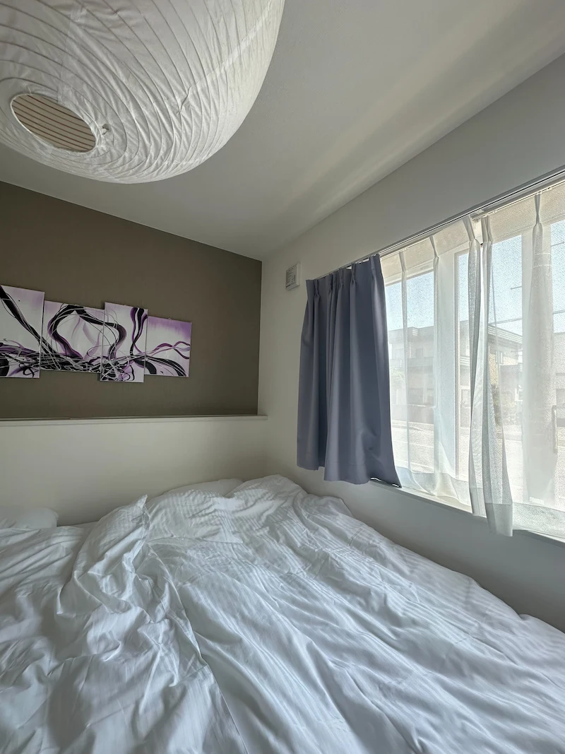A cozy, sunlit bedroom with an unmade white bed, purple curtains, and a large paper lantern light fixture.