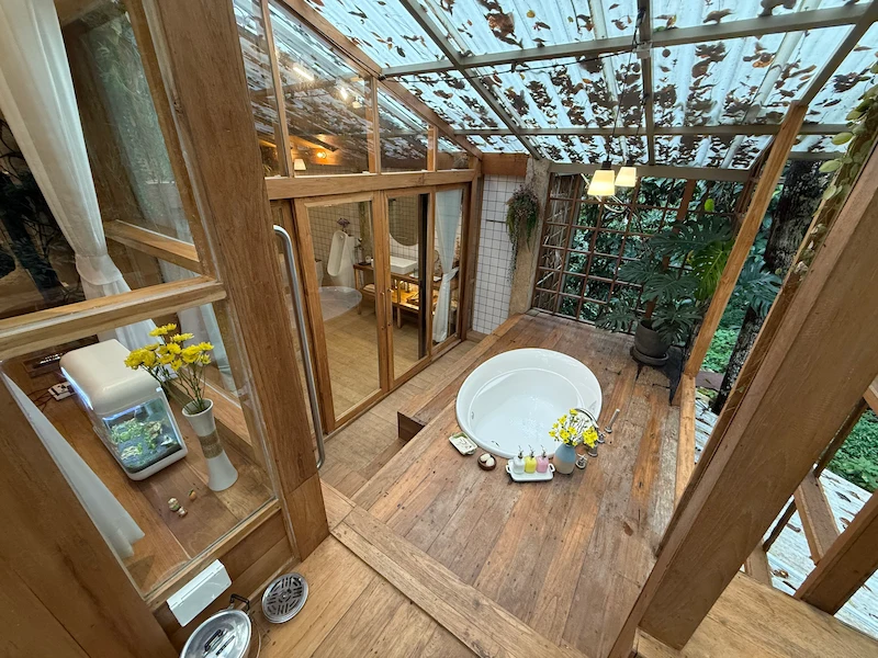 A luxurious white outdoor bathtub with jacuzzi functions located on a private wooden deck, surrounded by glass walls and forest views.
