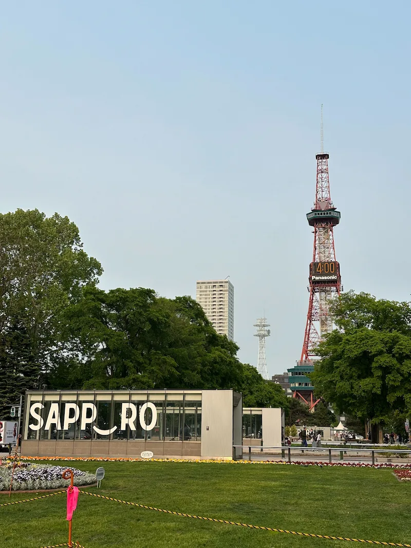 The iconic red Sapporo TV Tower rising above Odori Park, framed by a glass structure spelling out "SAPPORO" in the foreground.