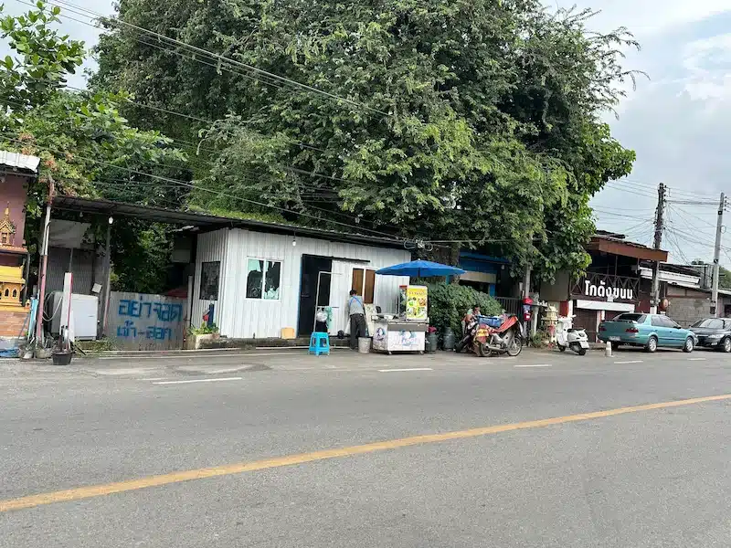 The roadside Khao Man Gai stall located next to an abandoned-style structure, featuring a food cart and small seating area under a blue umbrella.