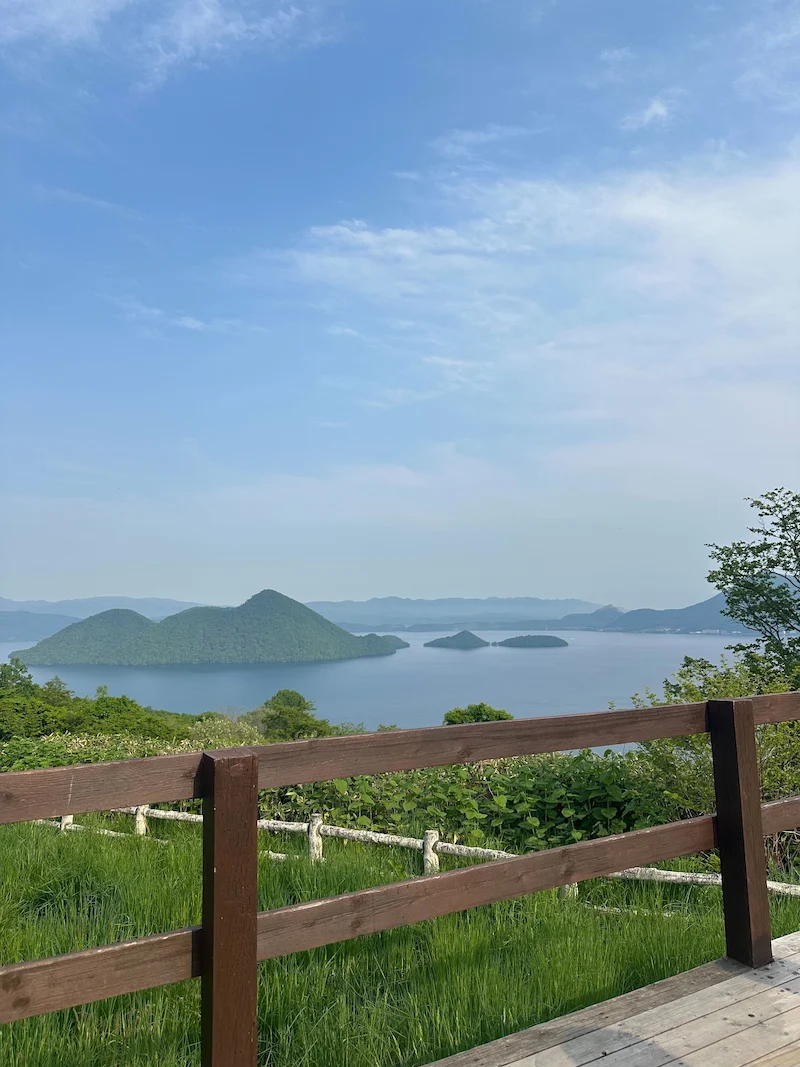 A scenic lookout point featuring a wooden fence and a panoramic view of the calm blue waters and islands of Lake Toya.