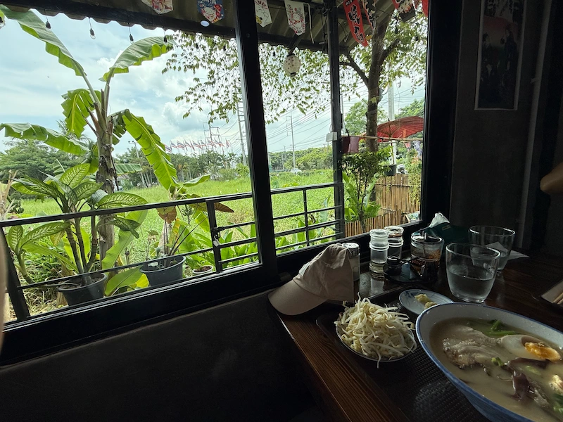 The view from inside Kinsou Ramen restaurant looking out a large window at a green field and banana trees, with a baseball cap resting on the table.