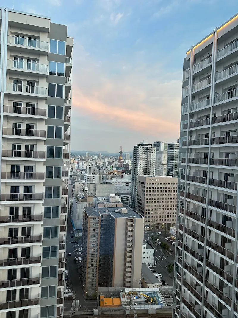 A high-rise view of the Sapporo city skyline and apartment buildings during sunset, with the TV Tower visible in the distance.