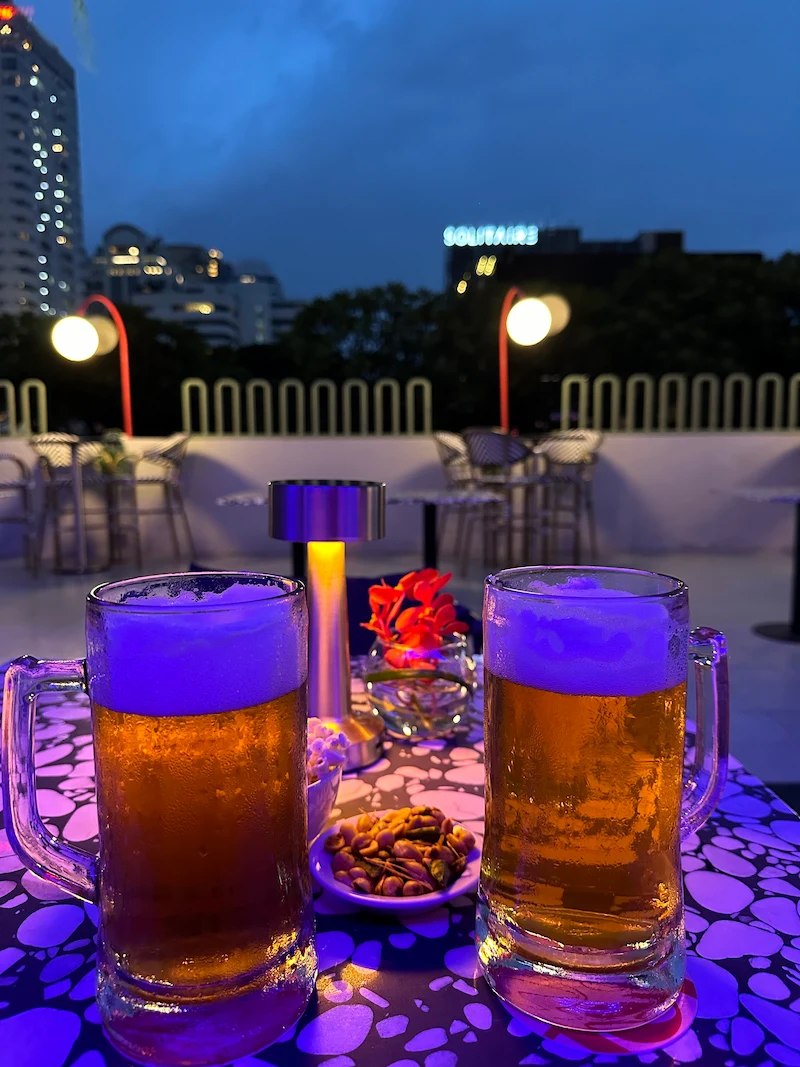 Two large, frosted mugs of cold beer sitting on a colorful table at an outdoor rooftop bar with city lights visible in the background.