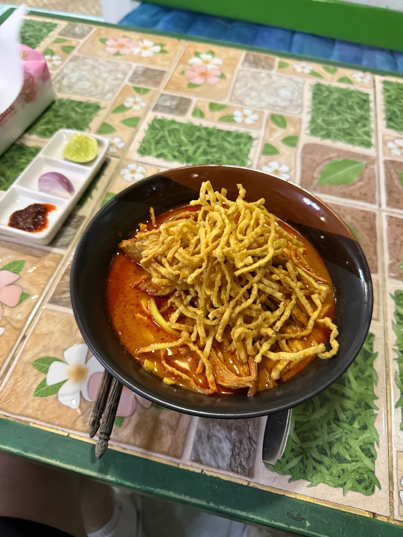 A bowl of northern Thai Khao Soi curry noodles topped with crispy fried noodles, served with a side of lime, red onion, and chili paste on a patterned tiled table.