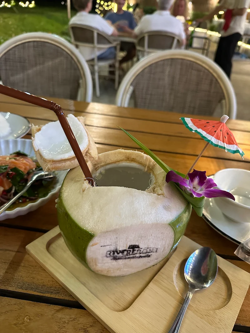 A fresh chilled young coconut served as a drink on a wooden tray, beautifully garnished with a purple orchid and a small colorful paper umbrella.