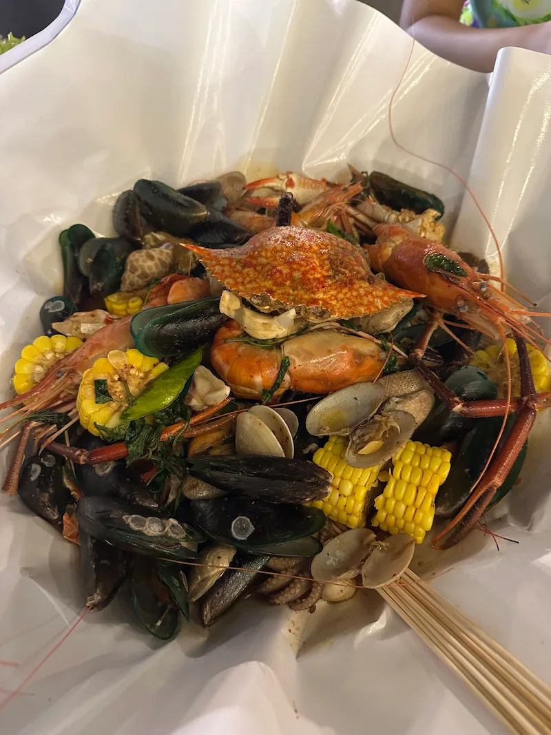 A large, steaming seafood boil piled high with grilled crab, giant prawns, mussels, clams, and corn on the cob served on white paper.