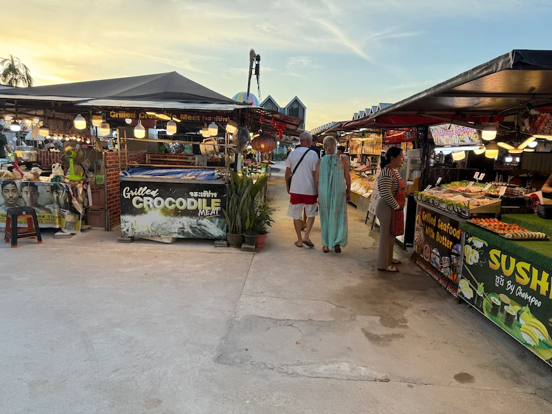 An outdoor Thai street food market at sunset featuring various stalls, including one prominently advertising "Grilled Crocodile Meat" as travelers walk through.