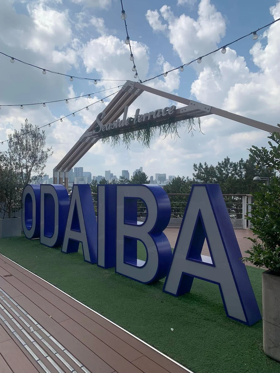 Large blue "ODAIBA" letters at the Seaside Terrace overlooking the Tokyo city skyline.