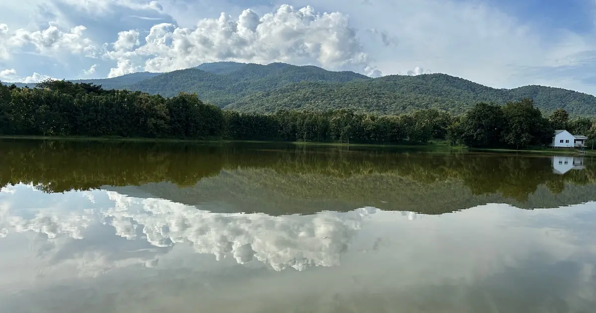 A serene view of a calm lake in Chiang Mai reflecting lush green mountains and white clouds under a bright blue sky.