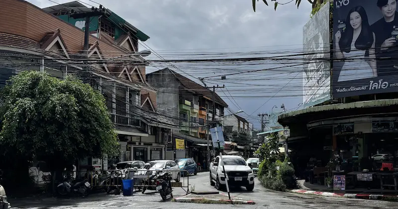 A busy local street in Chiang Mai featuring tangled overhead power lines, Thai storefronts, and traffic including a white pickup truck and parked scooters.