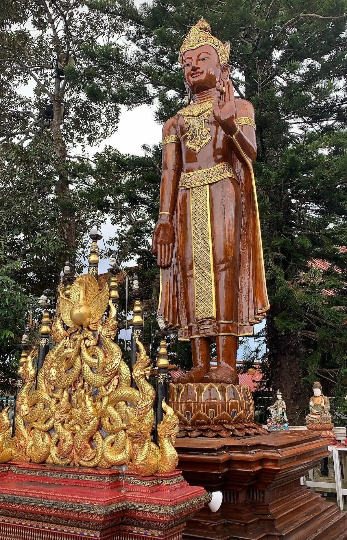 A tall, polished dark wood standing Buddha statue with golden adornments, situated behind intricate golden naga dragon sculptures at a Thai temple.