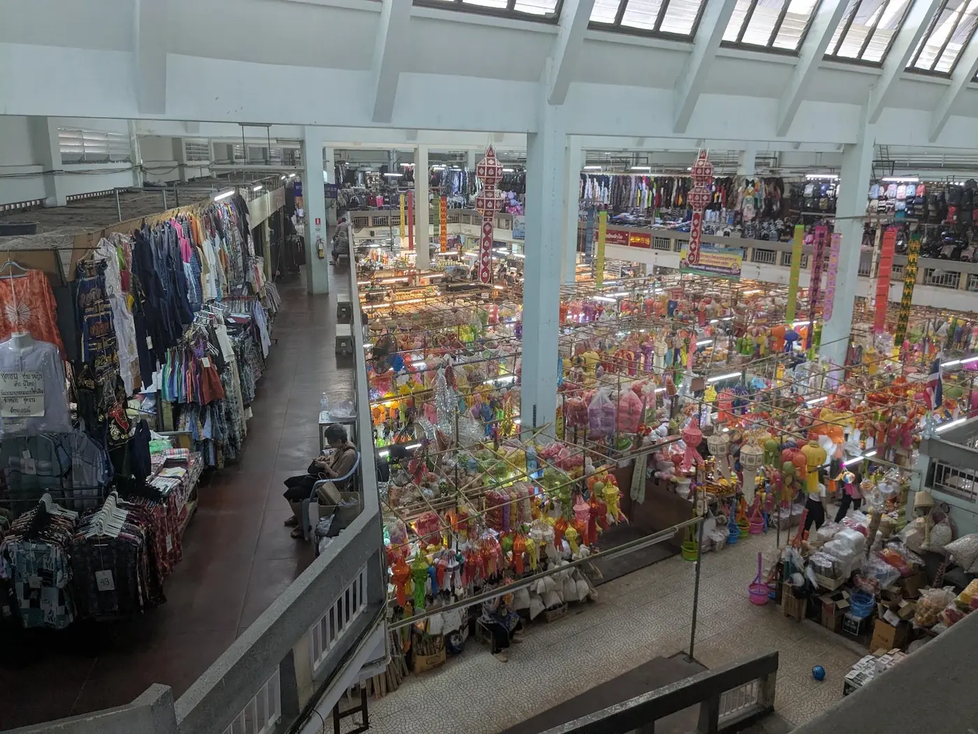 A high-angle view of the bustling interior of a large indoor market in Chiang Mai, showing rows of stalls selling clothes and colorful local decorations.