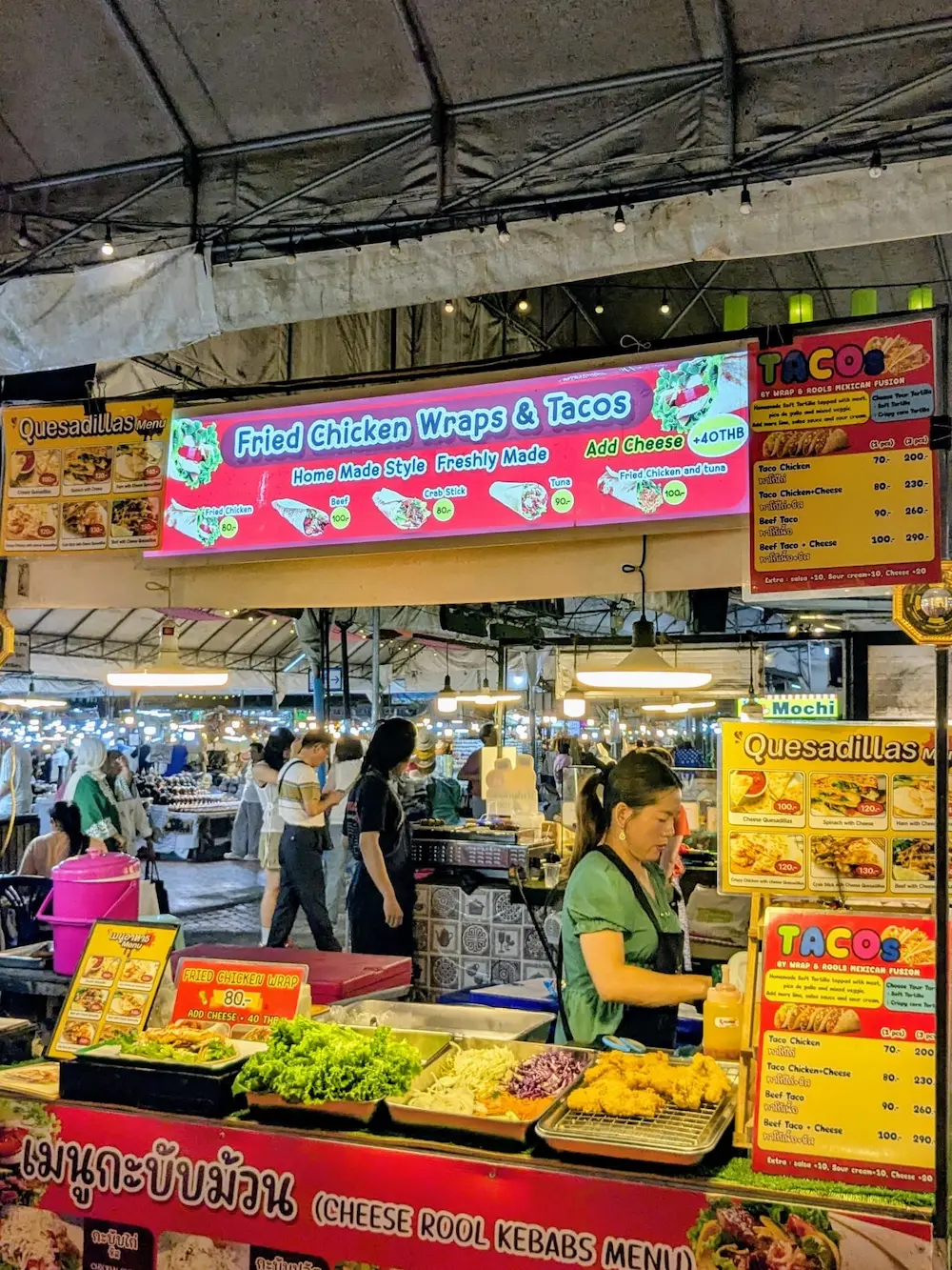 A brightly lit night market food stall in Chiang Mai selling fried chicken wraps and tacos, with a vendor working behind the counter.