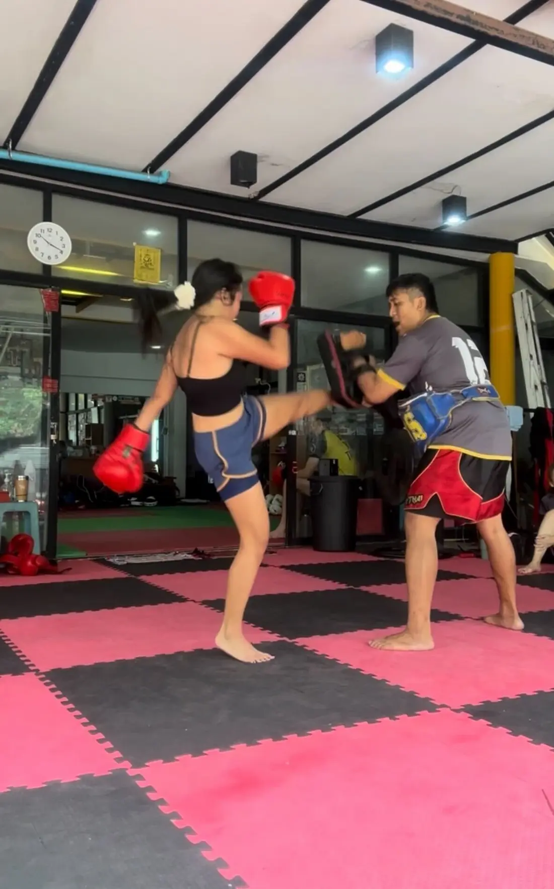A woman in a gym practicing Muay Thai, delivering a high roundhouse kick to a trainer’s pads while wearing red boxing gloves.