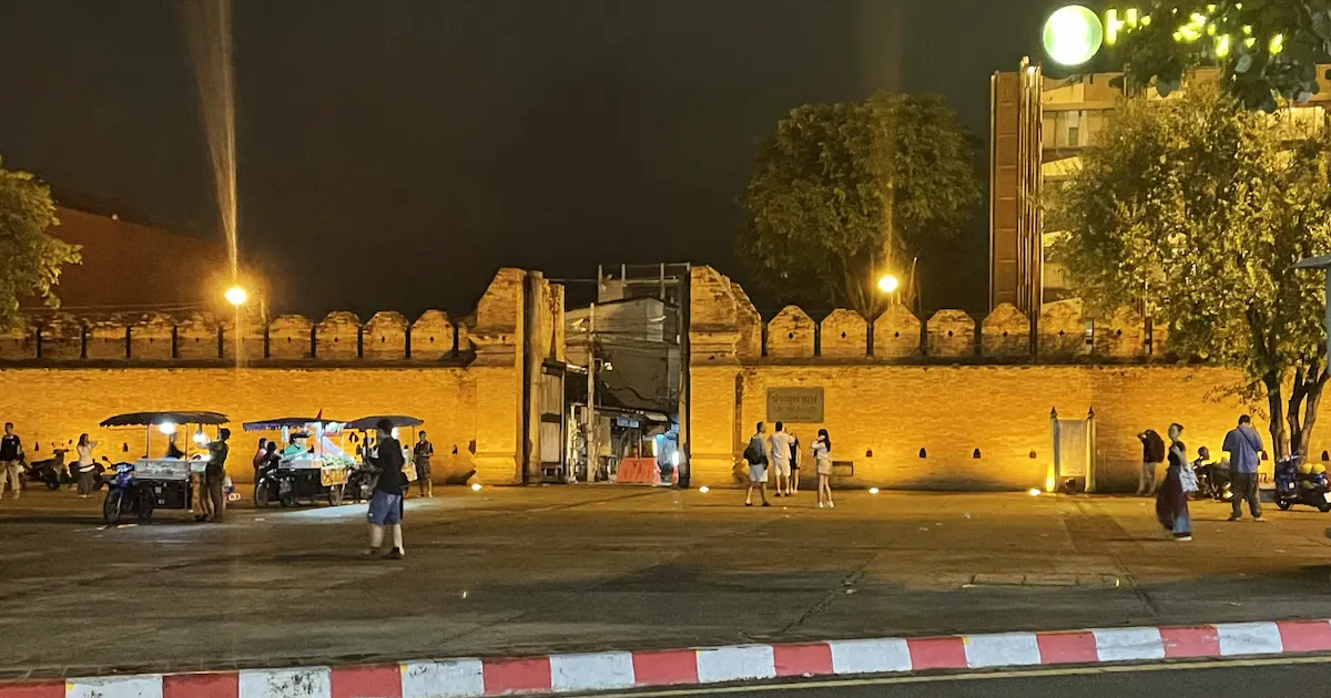 The historic red brick Thapae Gate in Chiang Mai at night, featuring street food vendors and tourists in the open plaza.