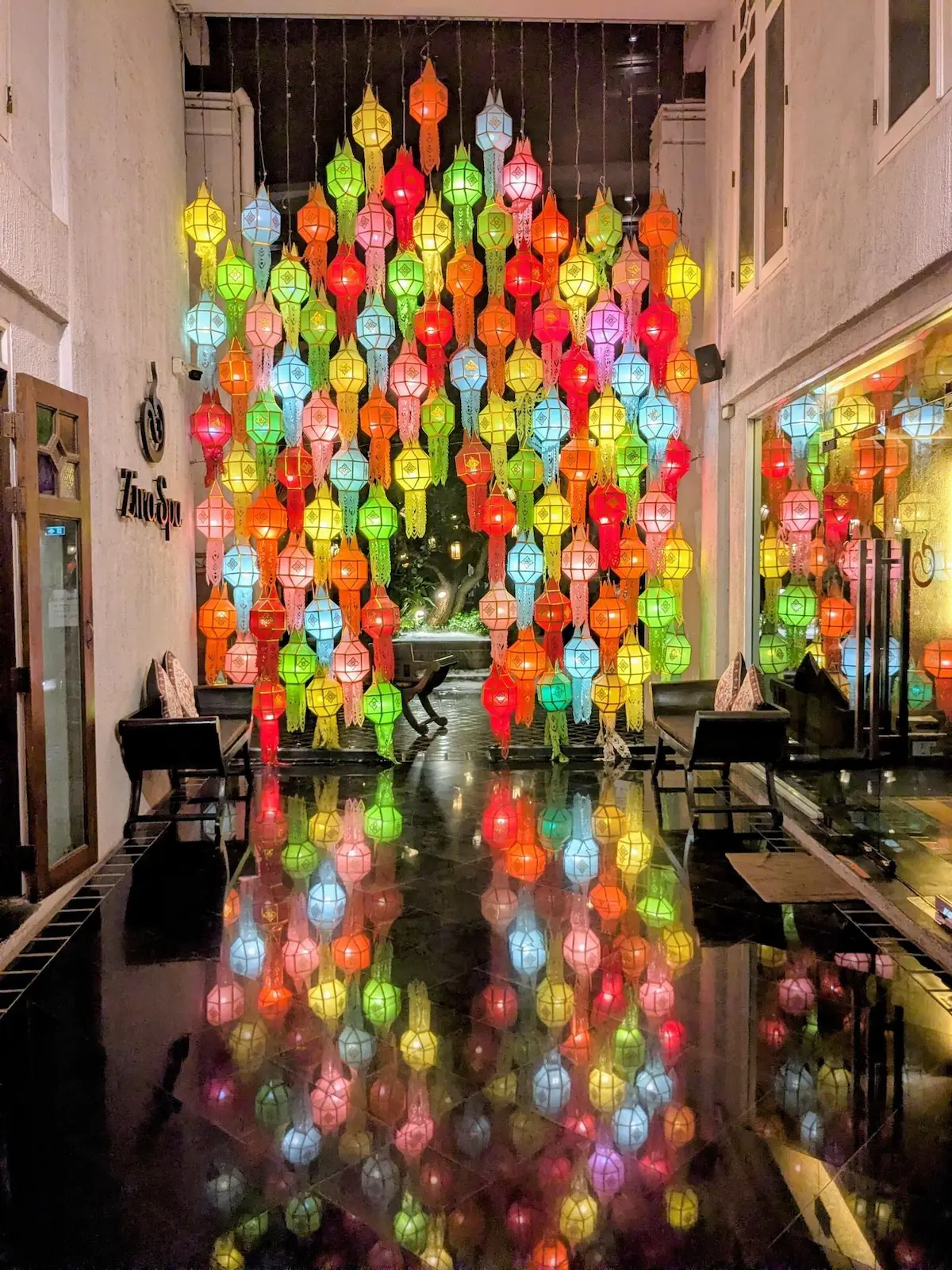 A vibrant display of colorful lit Lanna lanterns hanging in a hallway, their bright colors perfectly reflected on a polished dark floor.