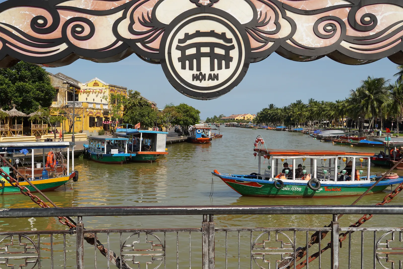 View of tourist boats and yellow riverside buildings in Hoi An through the ornate Hoi An bridge sign