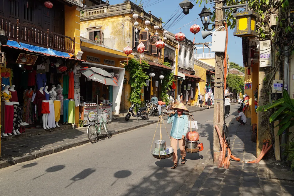 Street vendor carrying baskets on a shoulder pole past yellow colonial buildings and red lanterns in Hoi An