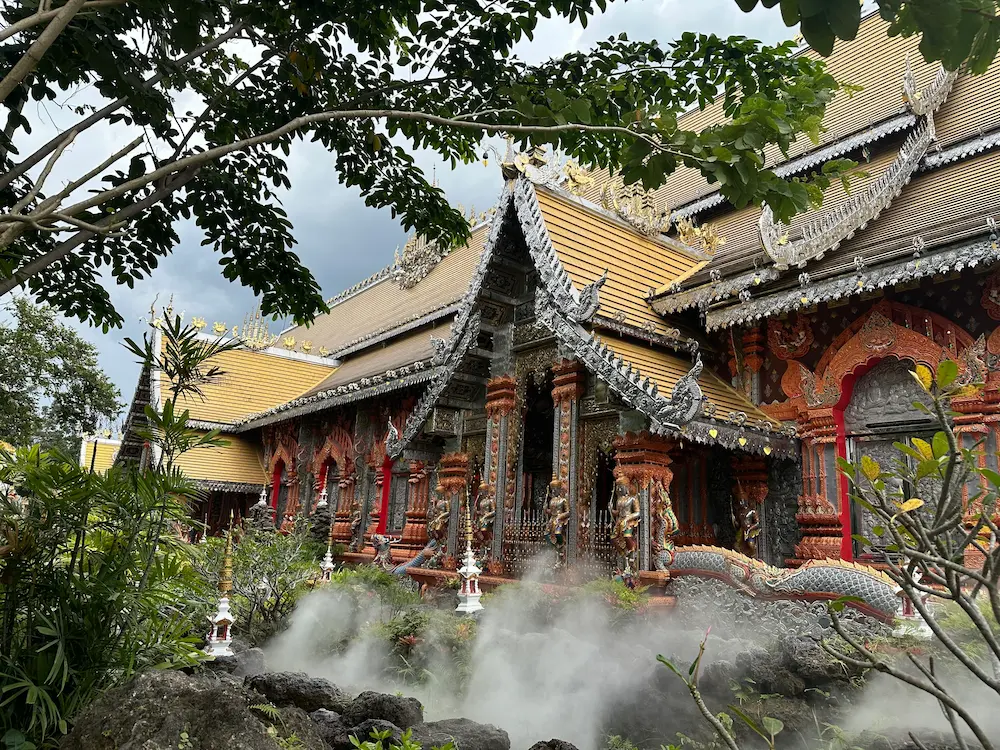 Silver and gold Lanna-style hall at Wat Sri Don Moon, Chiang Mai, with mist drifting over a rock garden and naga sculpture