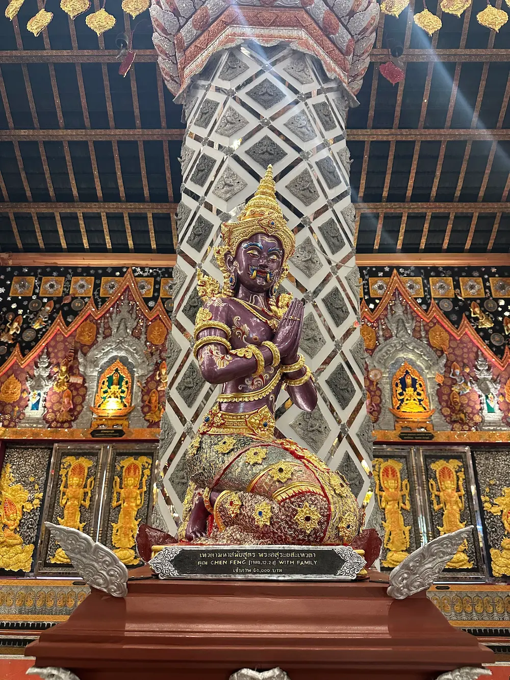 Purple kneeling guardian deity with a golden crown and hands in prayer, in front of a mirror-tiled pillar at Wat Sri Don Moon
